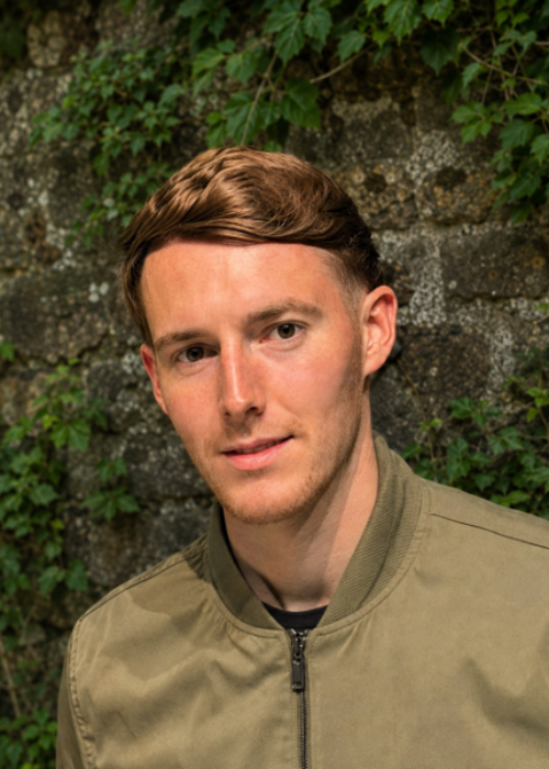 Headshot of man looking towards camera, against stone wall with some ivy.